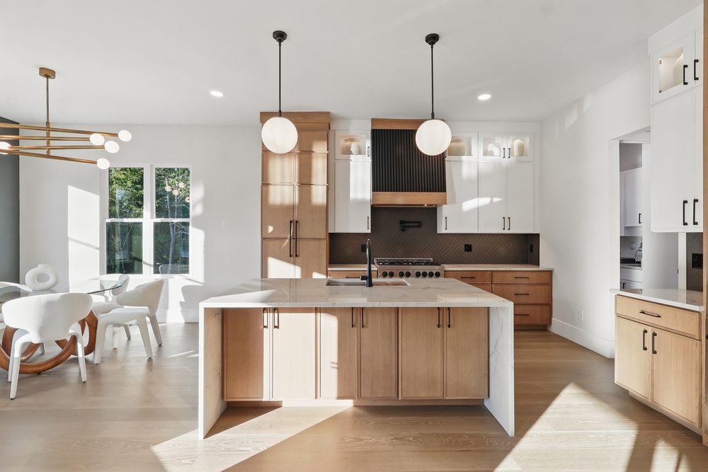 Two-tone kitchen with white upper cabinets, natural oak bases and a waterfall quartz island, looking toward the dining area